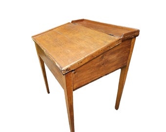 May include: A vintage wooden desk with a slanted writing surface and a small storage compartment. The desk is made of light brown wood and has four slender legs. The writing surface is approximately 24 inches wide and 18 inches deep.