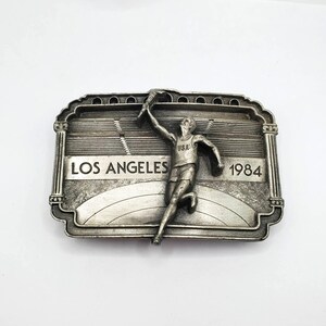 May include: Silver-toned belt buckle featuring a runner holding a torch, with "LOS ANGELES 1984" text. The buckle has a rectangular shape with an ornate border and a stadium design in the background. The runner is wearing a "USA" tank top.