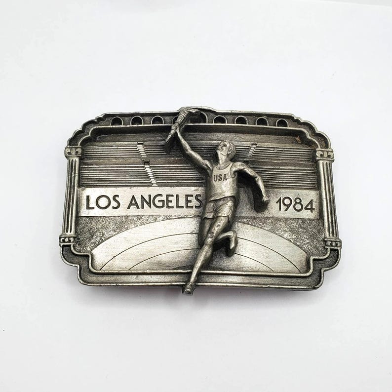 May include: Silver-toned belt buckle featuring a runner holding a torch, with "LOS ANGELES 1984" text. The buckle has a rectangular shape with an ornate border and a stadium design in the background. The runner is wearing a "USA" tank top.