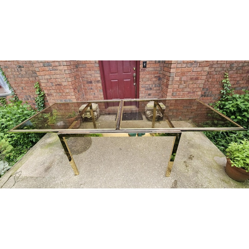 May include: A rectangular glass-top table with a gold-colored metal frame. The table has a two-piece glass top and is supported by four gold-colored legs. The table is outdoors, in front of a brick building.