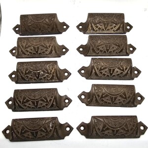 May include: A set of ten dark brown metal drawer pulls arranged in two columns. Each pull features a semi-circular design with embossed sunburst patterns, geometric lines, and small floral accents. Each piece has two screw holes for mounting.