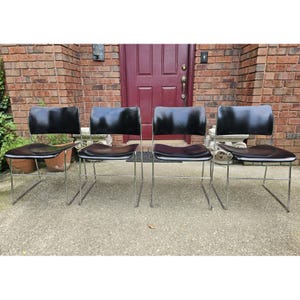May include: Four black chairs with chrome legs are arranged in a row. The chairs have a sleek, modern design with curved backs and seats. The chairs are set on a concrete surface in front of a brick building.