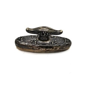 May include: A silver-toned, ornate, decorative, oval-shaped,  stamp pad with a detailed floral design and a  decorative handle.