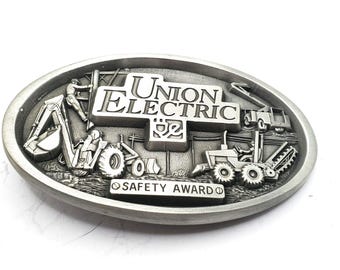 May include: An oval silver-toned metal belt buckle featuring a raised relief design of construction machinery and utility workers. The center displays the text "UNION ELECTRIC" with a power plug icon, and a banner below reads "SAFETY AWARD".