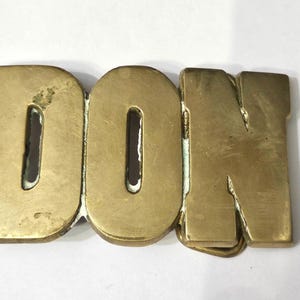 May include: A brass belt buckle featuring the letters D, O, and N connected together in a block font. The metal surface has a polished gold-toned finish with visible signs of wear and slight oxidation along the edges of the letters.