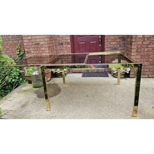May include: A rectangular coffee table with a clear glass top and a gold-colored metal frame. The table has a sleek, modern design with four square legs. The table is placed on a concrete surface in front of a brick wall.