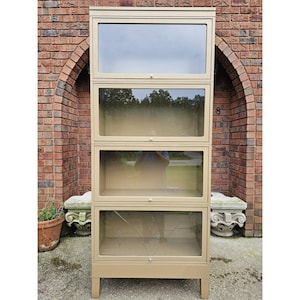 May include: A tall, beige metal bookcase with four glass-fronted compartments. The bookcase has a rectangular shape and sits on short legs. The glass panels reflect the surrounding environment. The bookcase is placed outside against a brick wall.