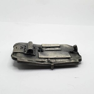 May include: A silver-toned metal belt buckle with a rectangular shape. The buckle features raised text and decorative elements, including a central cylindrical piece and several rectangular plaques with text. The buckle is on a white background.