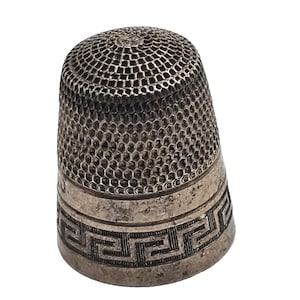May include: A silver-toned metal thimble featuring a textured, honeycomb-patterned top section and a smooth lower band decorated with a repeating geometric Greek key design. The surface shows signs of age and oxidation throughout the metal.