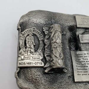 May include: A silver-toned metal belt buckle from the Arroyo Grande Buckle Company. The buckle features a relief of a figure, the text "Arroyo Grande", and the phone number 805/481-0719. The buckle has a textured background and a relief of a statue.
