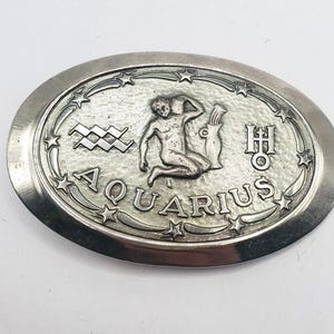 May include: An oval silver-toned metal belt buckle featuring a raised relief of the Aquarius zodiac figure pouring water from a vessel. The design includes the word AQUARIUS, a zigzag water symbol, an astrological glyph, and a border of small stars.
