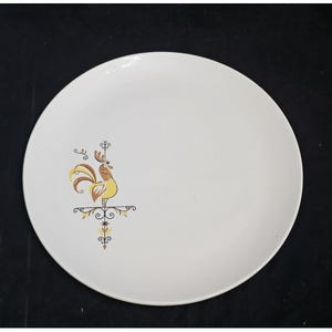 May include: A white ceramic dinner plate with a brown and yellow rooster design. The rooster is standing on a decorative brown and black metal stand.