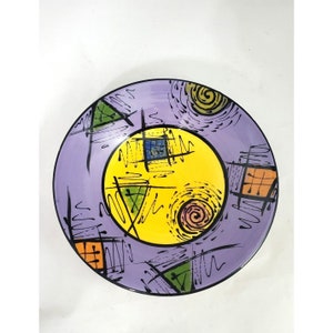 May include: A purple ceramic plate with a yellow center circle and black, green, and orange abstract designs.