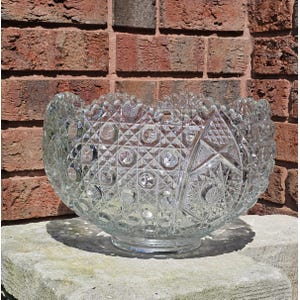 May include: A clear, cut-glass punch bowl with intricate geometric patterns. The bowl features a wide, scalloped rim and sits on a short, circular base. The cut glass design includes diamond, circle, and star shapes, creating a sparkling effect.