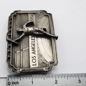 May include: A silver-toned metal souvenir from the 1984 Los Angeles Olympics. The rectangular piece features a stylized athlete, the text "LOS ANGELES" and "1984". The piece measures approximately 3 inches in length.