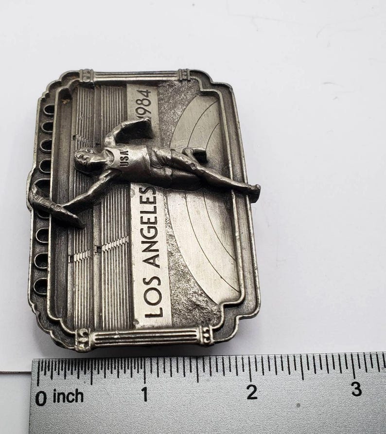 May include: A silver-toned metal souvenir from the 1984 Los Angeles Olympics. The rectangular piece features a stylized athlete, the text "LOS ANGELES" and "1984". The piece measures approximately 3 inches in length.