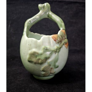 May include: A green ceramic vase with a twig handle and oak leaf design. The vase has a rounded shape and a textured surface.