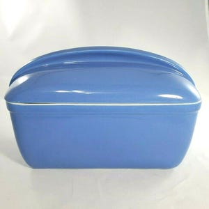 May include: A rectangular, glossy, cornflower blue ceramic bread box with a matching lid. The lid features a smooth, arched handle running along the top center. The container has rounded corners and a clean, solid-colored finish throughout.