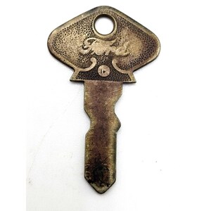 May include: Vintage Ford key with a brass finish. The key has a distinctive shape with the Ford logo embossed on the head. The key shows signs of wear, suggesting it is an antique or a used item. The key has a hole at the top for attachment.