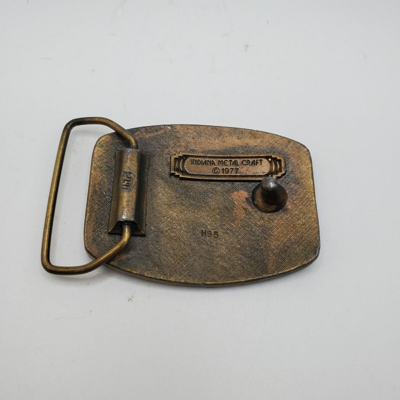 Op de afbeelding: Een messing gesp met een reli&euml;fdesign. De gesp heeft een metalen lus aan de achterkant en de tekst "INDIANA METAL CRAFT &copy; 1977" en "H95" aan de achterkant.