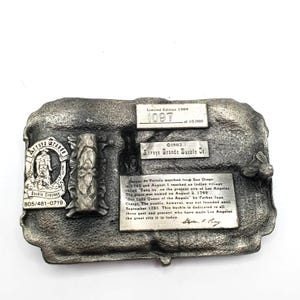 May include: A silver-toned metal belt buckle from Arroyo Grande Buckle Co. Features a relief of a figure, text, and the words "Limited Edition 1984". Includes the text "1097 of 10,000". The buckle commemorates the founding of Los Angeles.