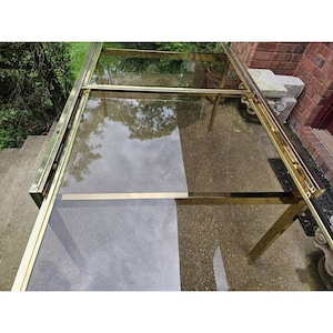 May include: A vintage brass and glass table with a rectangular glass top and a brass frame. The table has a reflective surface, and the brass has a warm, golden hue. The table is set on a concrete surface.