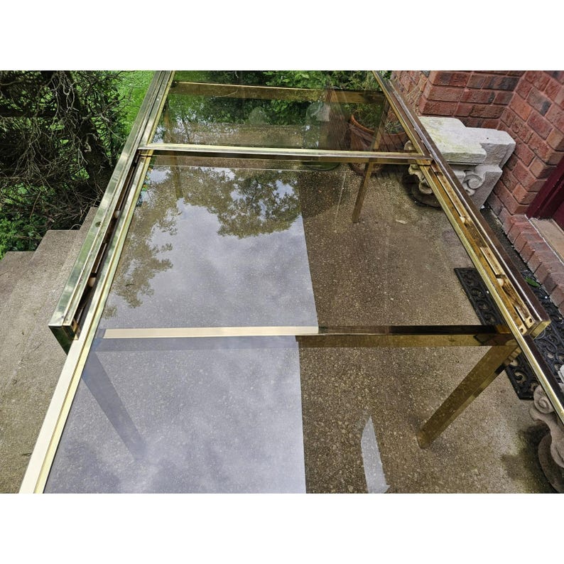 May include: A vintage brass and glass table with a rectangular glass top and a brass frame. The table has a reflective surface, and the brass has a warm, golden hue. The table is set on a concrete surface.