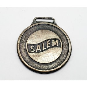 May include: A silver-toned metal watch fob with a black inscription that reads "SALEM" in a curved banner.