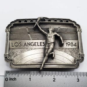May include: A silver-toned belt buckle featuring a runner holding a torch, with the text "LOS ANGELES 1984" and "USA". The buckle has a stadium design in the background and is approximately 3 inches wide.