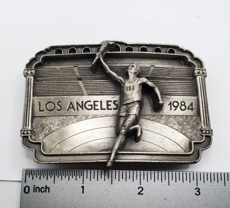 May include: A silver-toned belt buckle featuring a runner holding a torch, with the text "LOS ANGELES 1984" and "USA". The buckle has a stadium design in the background and is approximately 3 inches wide.