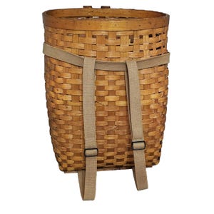 May include: A woven backpack basket made of light brown wood. The basket has a rectangular shape with rounded top edges. It features tan straps and black buckles for carrying. The basket is designed for outdoor use.
