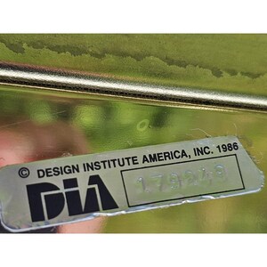 May include: A close-up of a metallic gold-colored object with a label. The label reads "DESIGN INSTITUTE AMERICA, INC. 1986" with the letters "DIA" in a stylized font and a serial number. The background is blurred.