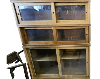 May include: A wooden bookcase with three sections, each with sliding glass doors. The bookcase is made of light-colored wood and has a rectangular shape. The doors are clear, allowing visibility into the shelves.