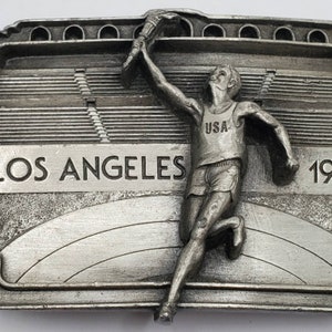 May include: A silver-colored metal belt buckle featuring a running athlete holding a torch aloft. The buckle is inscribed with "LOS ANGELES 1984" and the athlete wears a "USA" tank top. The background depicts a stadium.