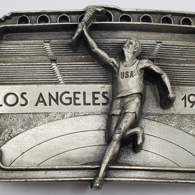 May include: A silver-colored metal belt buckle featuring a running athlete holding a torch aloft. The buckle is inscribed with "LOS ANGELES 1984" and the athlete wears a "USA" tank top. The background depicts a stadium.
