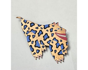 May include: A gold and blue painted wooden animal brooch with a toothy grin. The animal has a spotted pattern and a long tail.
