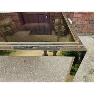 May include: A rectangular coffee table with a brass-colored metal frame and a clear glass top. The table has a simple, geometric design and is set outdoors on a concrete surface.