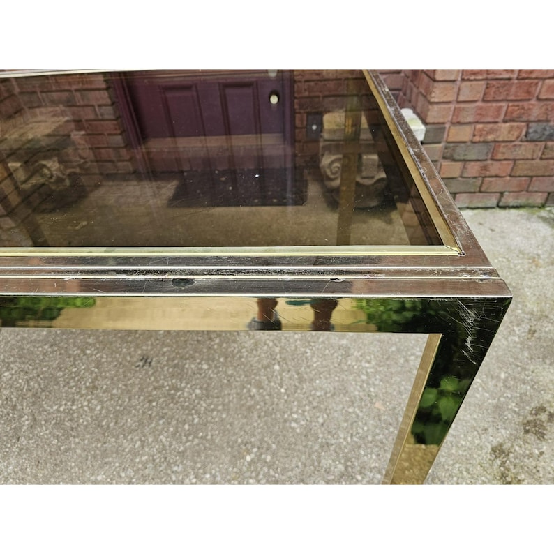 May include: A rectangular coffee table with a brass-colored metal frame and a clear glass top. The table has a simple, geometric design and is set outdoors on a concrete surface.