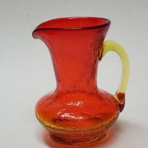 May include: A small, red glass pitcher with a yellow handle. The pitcher has a textured, crackled glass surface.