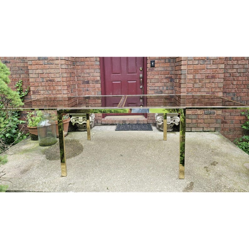 May include: A rectangular glass-top table with a brass-colored metal frame. The table has four brass-colored legs and is set on a concrete patio. The background includes a brick wall, a red door, and some greenery.