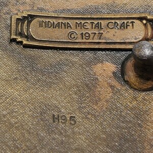 Op de afbeelding: Een close-up van een messing gesp met de tekst "Indiana Metal Craft &copy; 1977" en het nummer "H95" gegraveerd op het oppervlak.