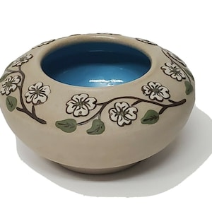 May include: A small, cream-colored ceramic bowl with a blue glaze inside. The bowl is decorated with a brown floral design featuring white flowers and green leaves.
