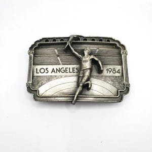 May include: A silver-toned metal belt buckle featuring a running figure holding a torch. The buckle is rectangular with the words "LOS ANGELES 1984" and a stadium design. The figure is wearing a USA tank top.