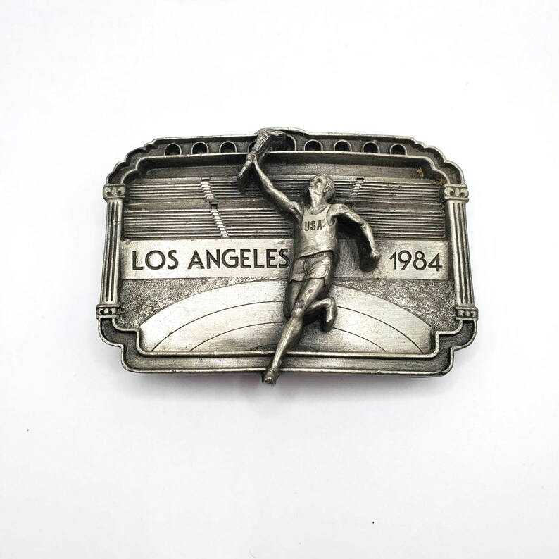 May include: A silver-toned metal belt buckle featuring a running figure holding a torch. The buckle is rectangular with the words "LOS ANGELES 1984" and a stadium design. The figure is wearing a USA tank top.