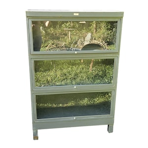May include: A three-tiered, gray-green display cabinet with glass doors. The cabinet has a rectangular shape and sits on short legs. The glass doors reflect the outdoor surroundings, including greenery and a person.