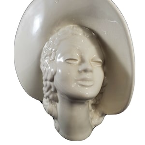 May include: Off-white ceramic wall hanging featuring a woman's head wearing a wide-brimmed hat. The sculpture has a smooth, glossy finish and detailed facial features, including closed eyes and defined lips. The hat's brim curves around the head, creating a classic, elegant look.