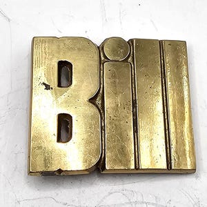 May include: A vintage brass sign spelling out the word "Bill". The letters are thick and blocky, with the "B" having two cut-out sections. The "i" is a vertical rectangle, and the two "l"s are also vertical rectangles. The sign has a worn, aged appearance.