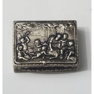 May include: A silver trinket box with an ornate design featuring a group of cherubs playing with a goat in a wooded setting.