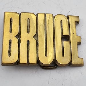 May include: A gold-colored metal belt buckle with the word "BRUCE" embossed on it.