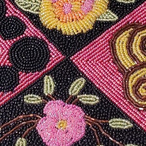 May include: Close-up of a beaded fabric with a floral design. The fabric features pink, yellow, and green beads arranged in a pattern of flowers and leaves. The background is black.